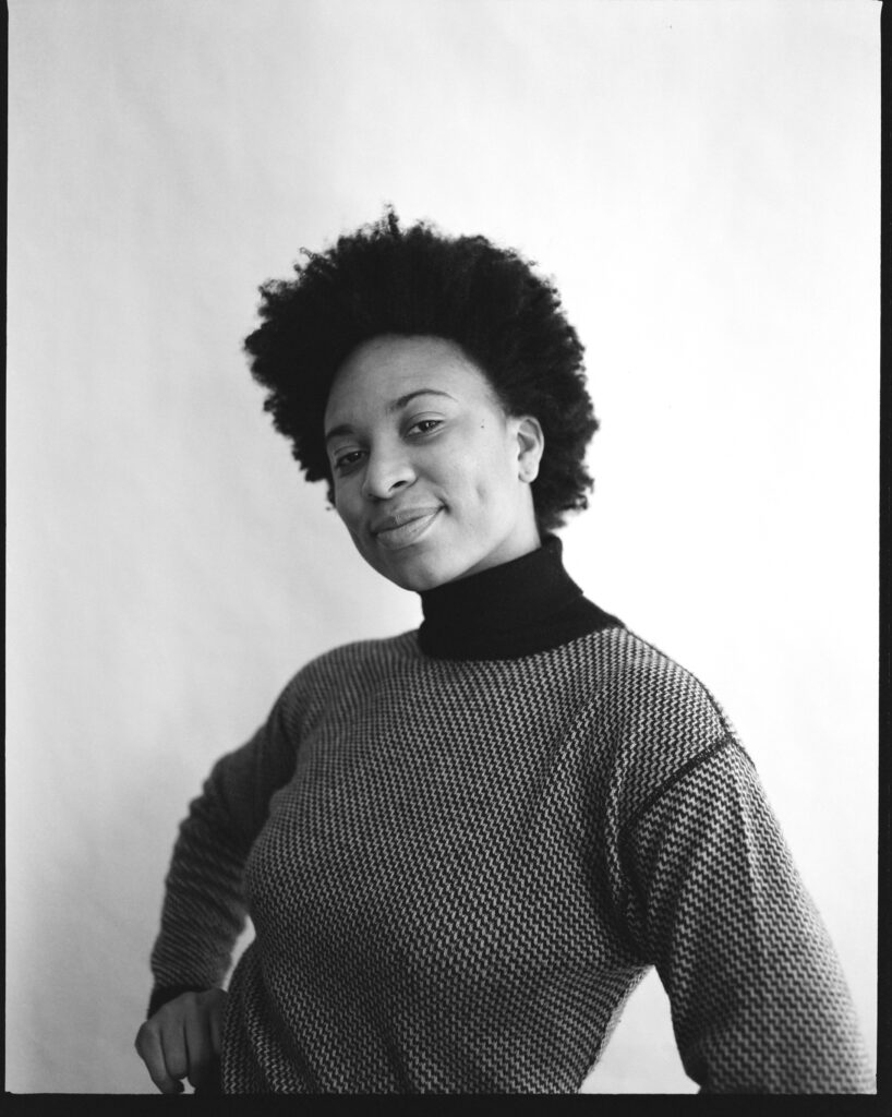 Black and white photograph of Kira Joy Williams smiling in a turtleneck with hands on hips