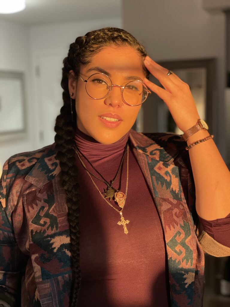 Portrait photograph of a fair-skinned person standing in golden hour sunlight. They're wearing glasses and a maroon turtle neck with jewelry. 