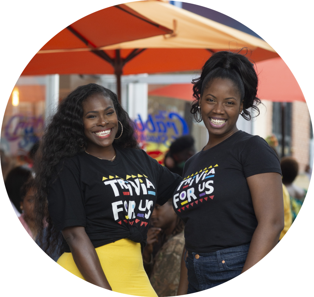 Two black femmes wearing black shirts with Trivia For Us's logo, smiling at the camera. 